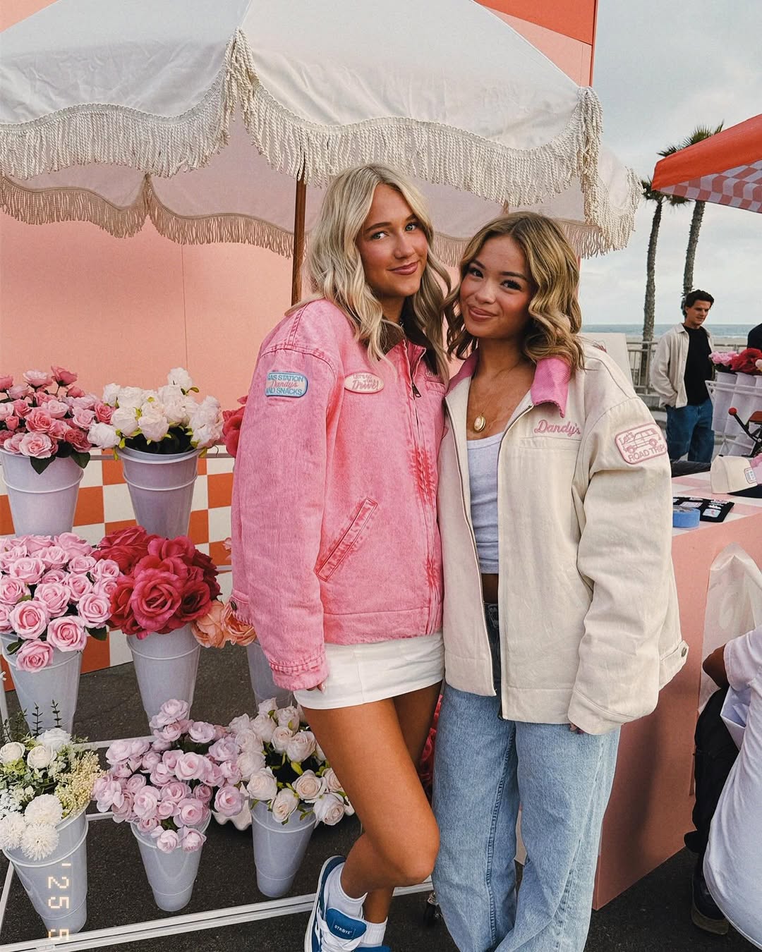 Two women stand smiling in front of pink floral displays and a white fringe umbrella. One wears a pink jacket and white skirt; the other wears a white jacket and jeans. The setting is festive and outdoors, with flowers and pastel decor.