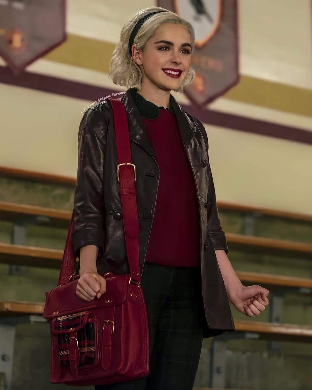 A young woman with platinum blonde hair, wearing a black headband, maroon top, and dark leather coat, stands smiling in bleachers with a large red satchel—a perfect 90s outfit inspo. Banners and wooden benches are visible in the background.