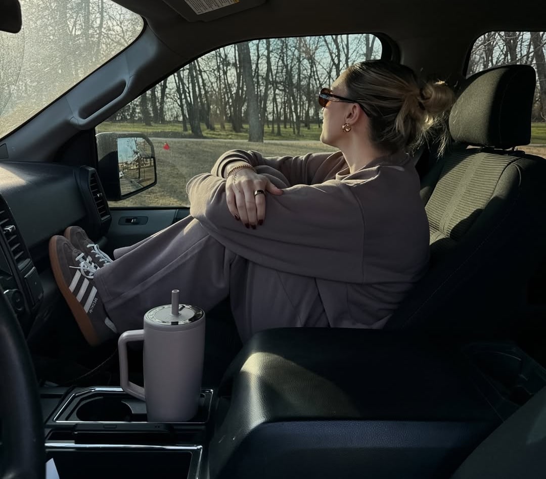 A woman in sunglasses and casual clothes sits in the front passenger seat of a car with her knees up, looking out the window at trees. Sunlight filters in as she enjoys quiet moments and contemplates new winter outfits or fresh outfit formulas.