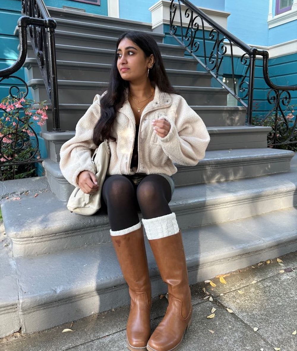 A woman with long dark hair sits on outdoor gray steps, showcasing winter fashion in a cream fleece jacket, black tights, brown knee-high boots with white leg warmers, and holding a light tote bag—perfect inspiration for reviving a tired wardrobe.