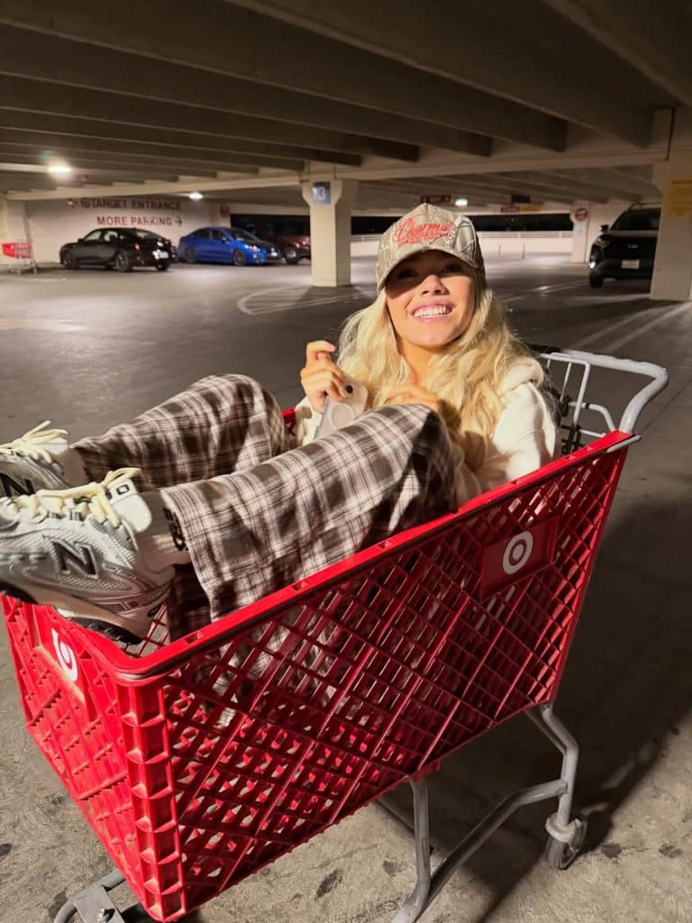 A smiling woman in plaid pants, sneakers, and a baseball cap sits inside a red Target shopping cart in a dimly lit parking garage.