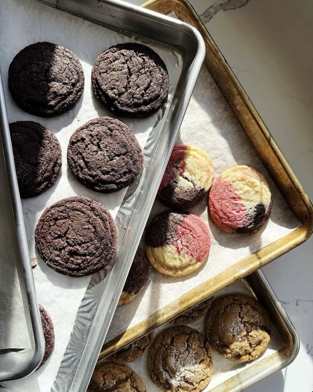 Three baking sheets with assorted cookies: dark chocolate, tri-colored (pink, yellow, brown), and golden brown cookies, all on parchment paper, in sunlight on a marble surface.