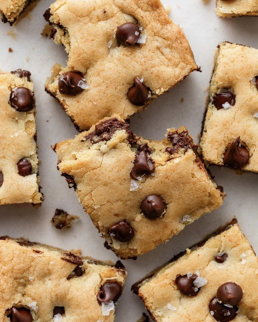 Top-down view of chocolate chip cookie bars cut into squares, some with bites taken out. The bars have a golden-brown crust, visible chocolate chips, and a light sprinkle of flaky salt on top.