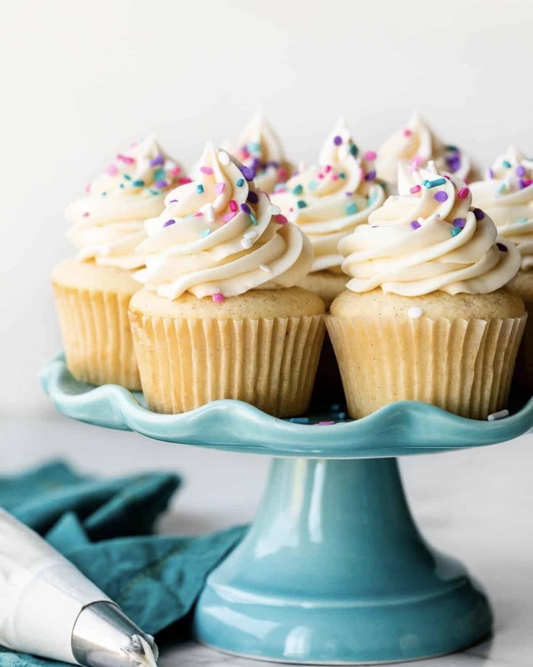 Six vanilla cupcakes with swirled white frosting and colorful sprinkles are displayed on a light blue cake stand. A piping bag and a teal napkin are beside the stand.