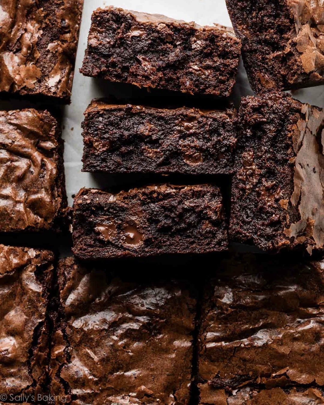 A close-up view of rich, fudgy chocolate brownies, some cut into squares and stacked on top of each other, showing a moist, dense texture and shiny, crackly tops.