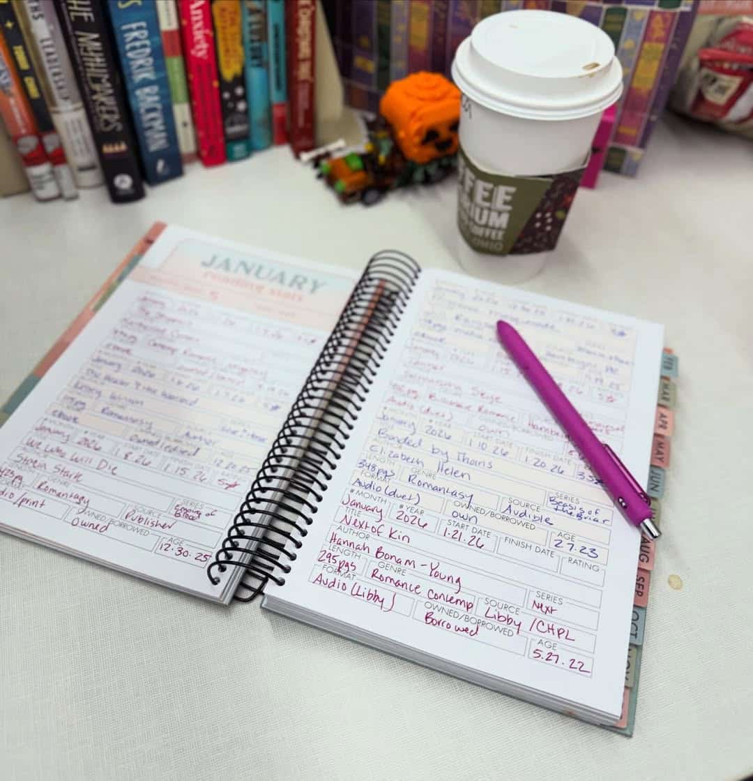 An open planner filled with handwritten notes sits on a table next to a purple pen and a coffee cup. Books are lined up in the background, creating a cozy, organized workspace.
