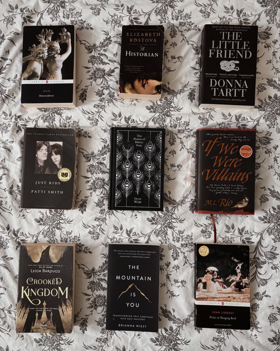 Nine books are arranged in a neat grid on a white floral-patterned bedspread, with covers facing up. Titles include The Song of Achilles, The Historian, The Little Friend, and others.