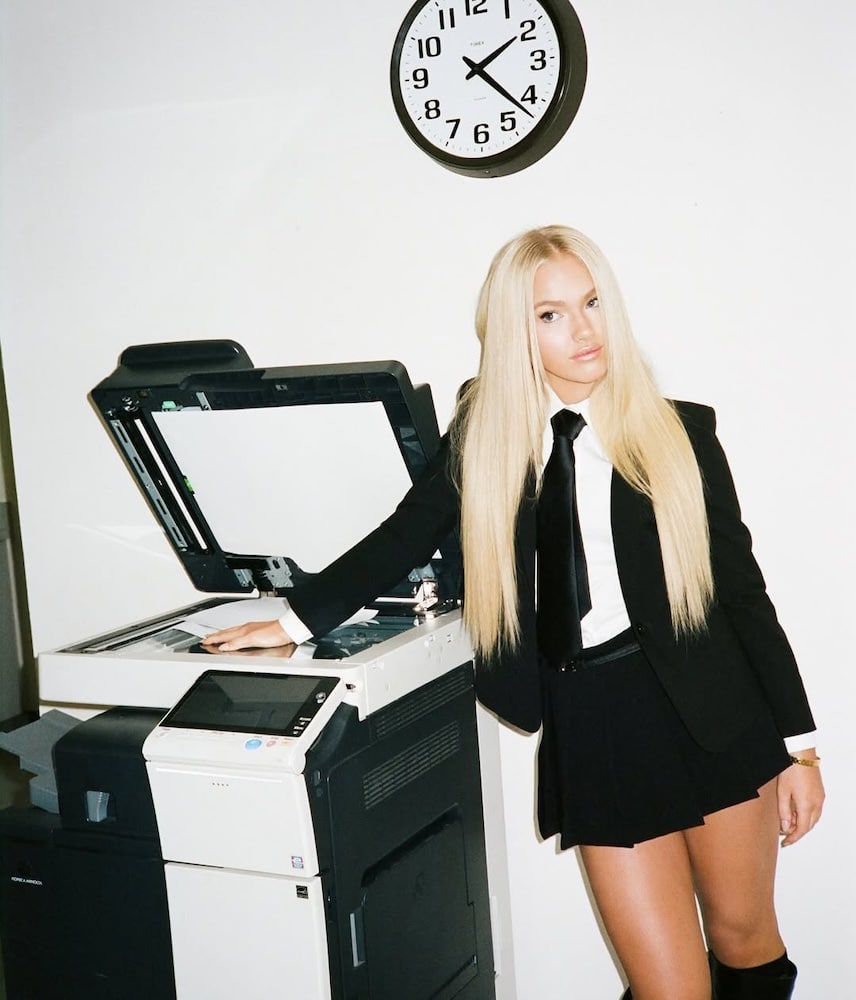 A woman with long blonde hair, channeling outfit inspo, wears a black blazer, white shirt, black tie, and skirt as she stands by a photocopier. A wall clock shows 11:10 in the office setting.