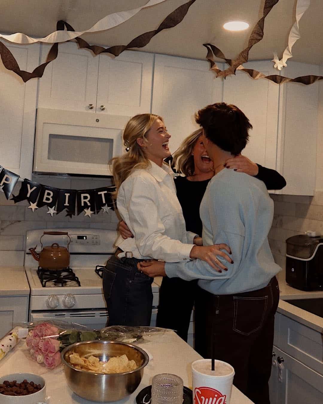 Three people are laughing and hugging in a decorated kitchen with a “HAPPY BIRTHDAY” banner. Snacks, flowers, and a Swig drink cup are on the counter—proof that friends who stay calm together can handle drama and celebrate joyfully.