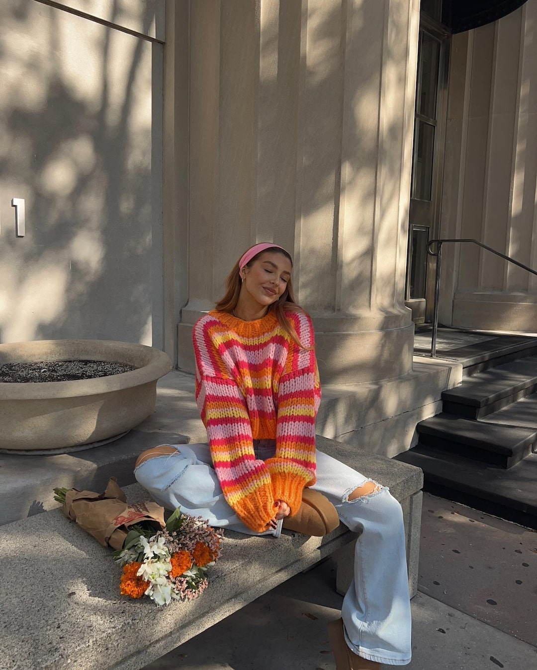 A woman sits on a concrete ledge outside, her spring wardrobe shining in a colorful orange and pink striped sweater, ripped light blue jeans, tan boots, and a pink headband. She smiles peacefully with eyes closed, next to a bouquet of flowers.