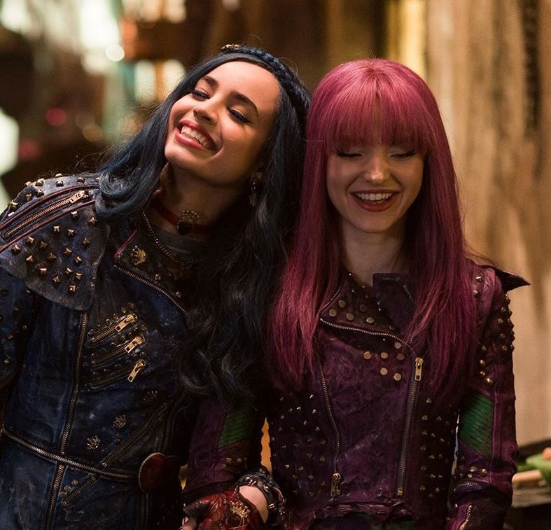 Two young women wearing studded jackets and colorful hair—one with long dark blue hair, the other with straight bright purple hair—stand close together, smiling and looking down.