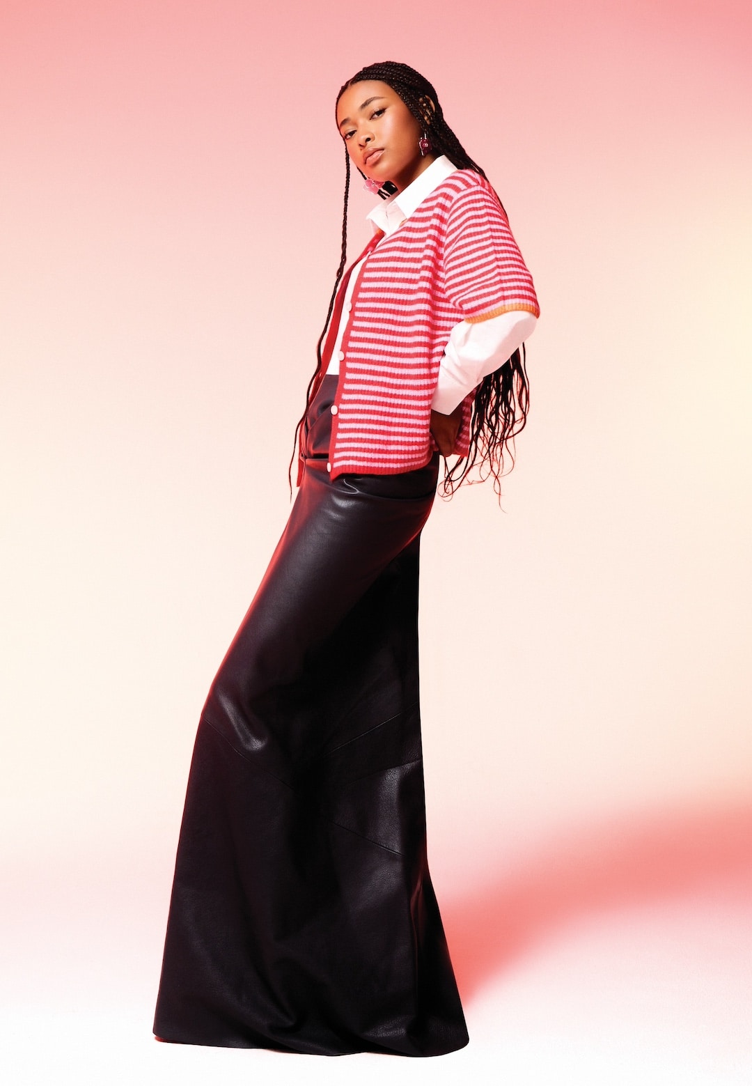 A woman poses confidently in a red and white striped short-sleeve cardigan over a white shirt with a long black skirt—a perfect spring outfit. The pink gradient background adds a modern, stylish vibe to this spring fit.