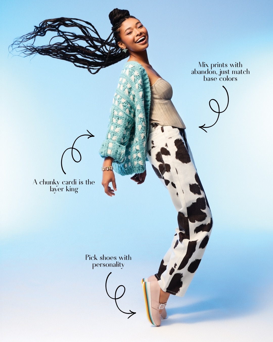 A smiling woman poses mid-jump against a blue background, wearing a chunky teal cardigan, beige top, cow-print pants, and rainbow platform shoes. This spring fashion style guide highlights mixing prints, layering cardigans, and choosing bold shoes.