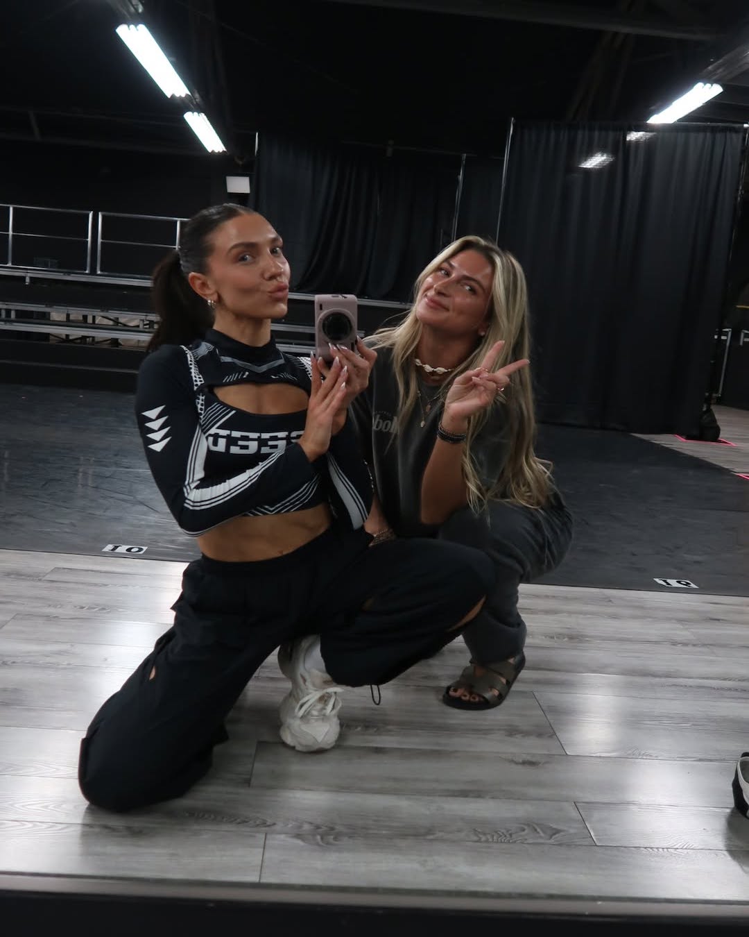 Two women pose in front of a large mirror on a light wood floor. One kneels holding a phone, the other crouches making a peace sign. Both are casually dressed, showing how to stay calm and collected even with a black curtain and stage setup in the background.