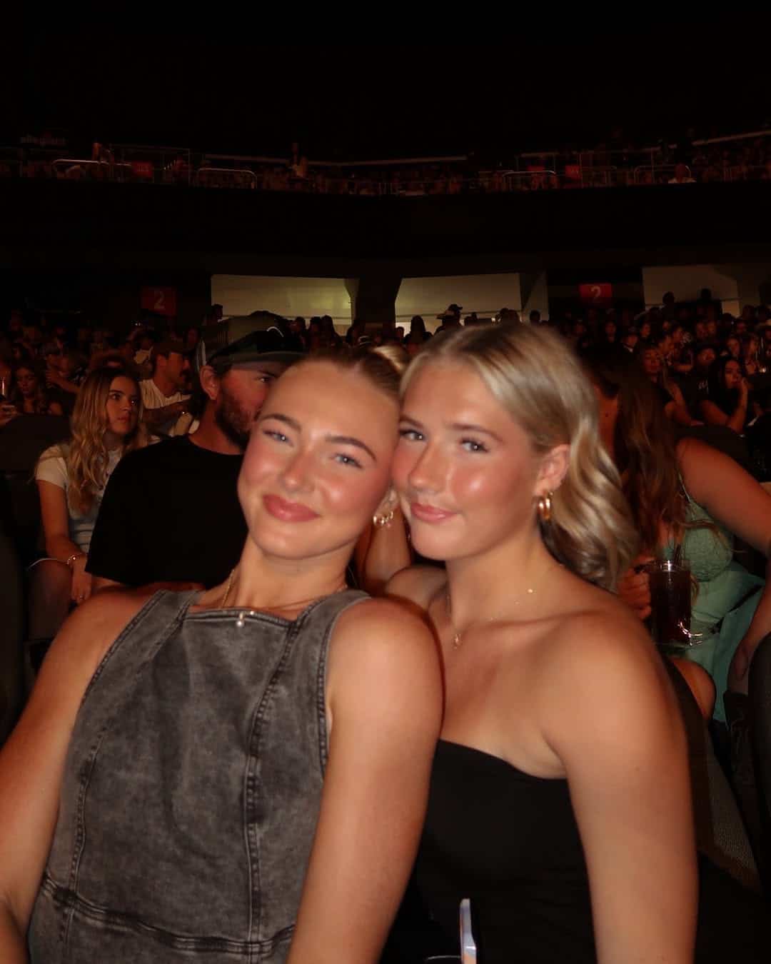 Two young women sit close together and smile at the camera in a crowded indoor arena, their warmth and happiness suggesting they know how to stay calm and handle drama, even amid the bustling crowd around them.