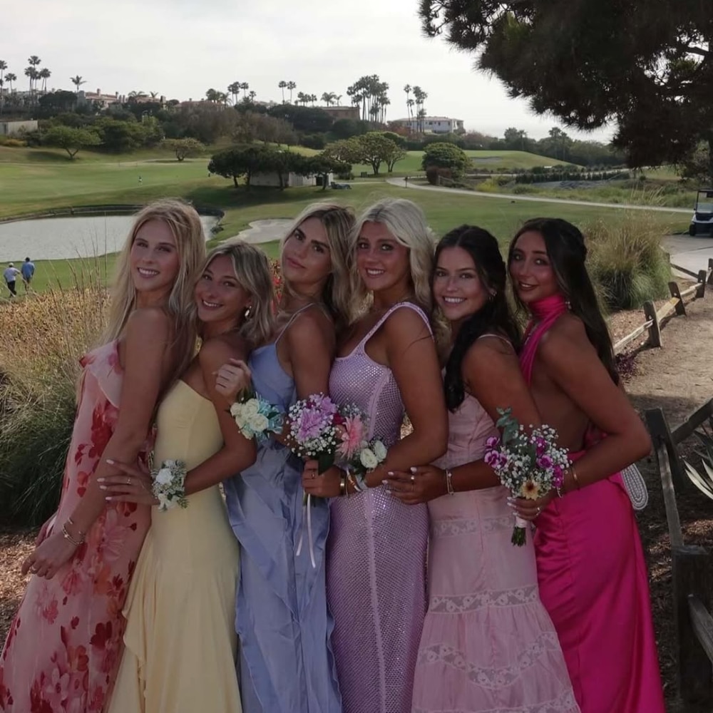 Six young women in colorful dresses stand outdoors in a line, holding small bouquets and smiling at the camera, with a golf course and trees in the background on a sunny day.