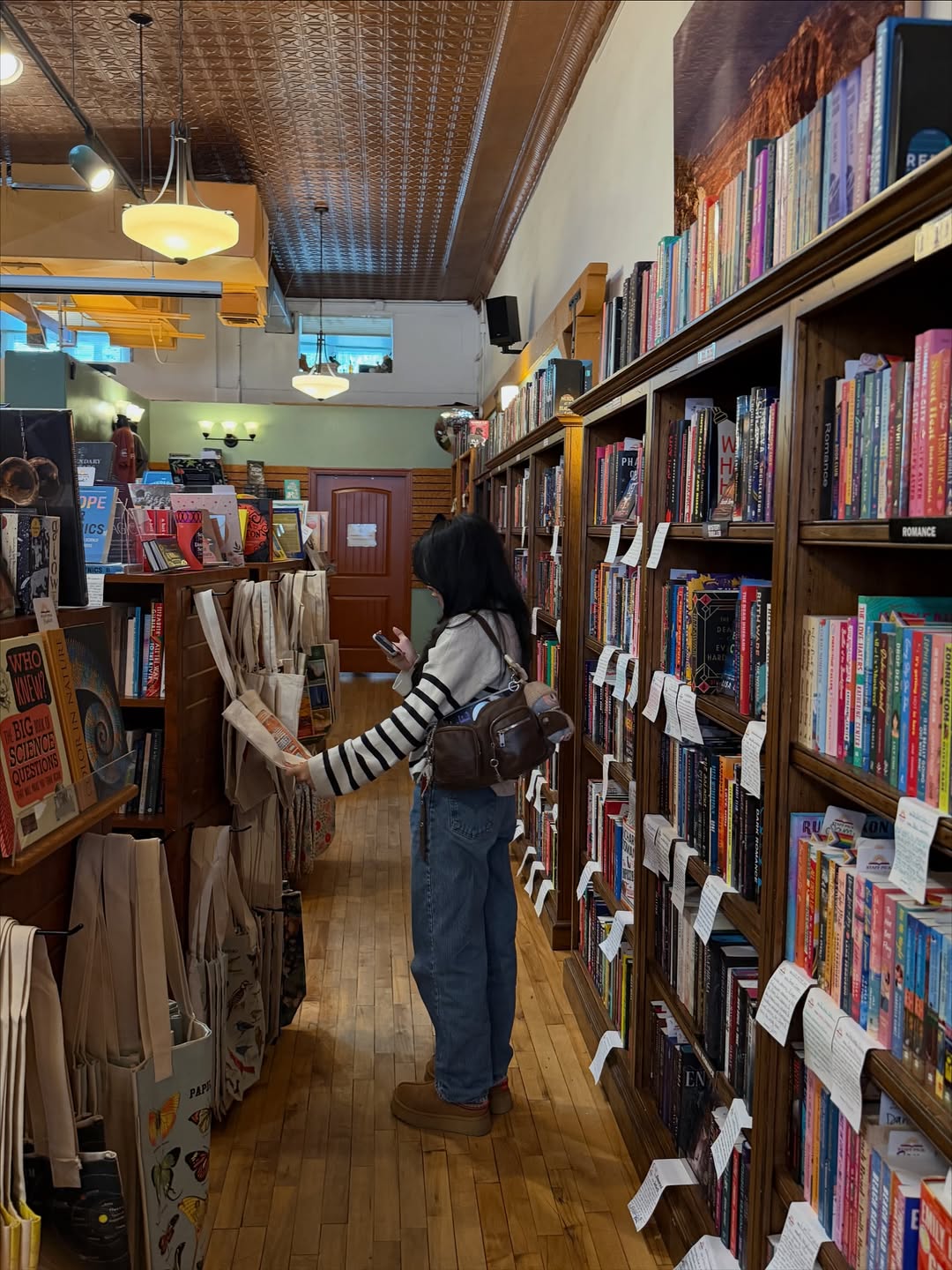 A person wearing a striped sweater, jeans, and a backpack browses Classic Reads in a cozy bookstore, holding a tote bag and phone. Bookshelves and book-filled tables line the wooden floor, with handwritten notes suggesting How To Get Into Classic Reads.