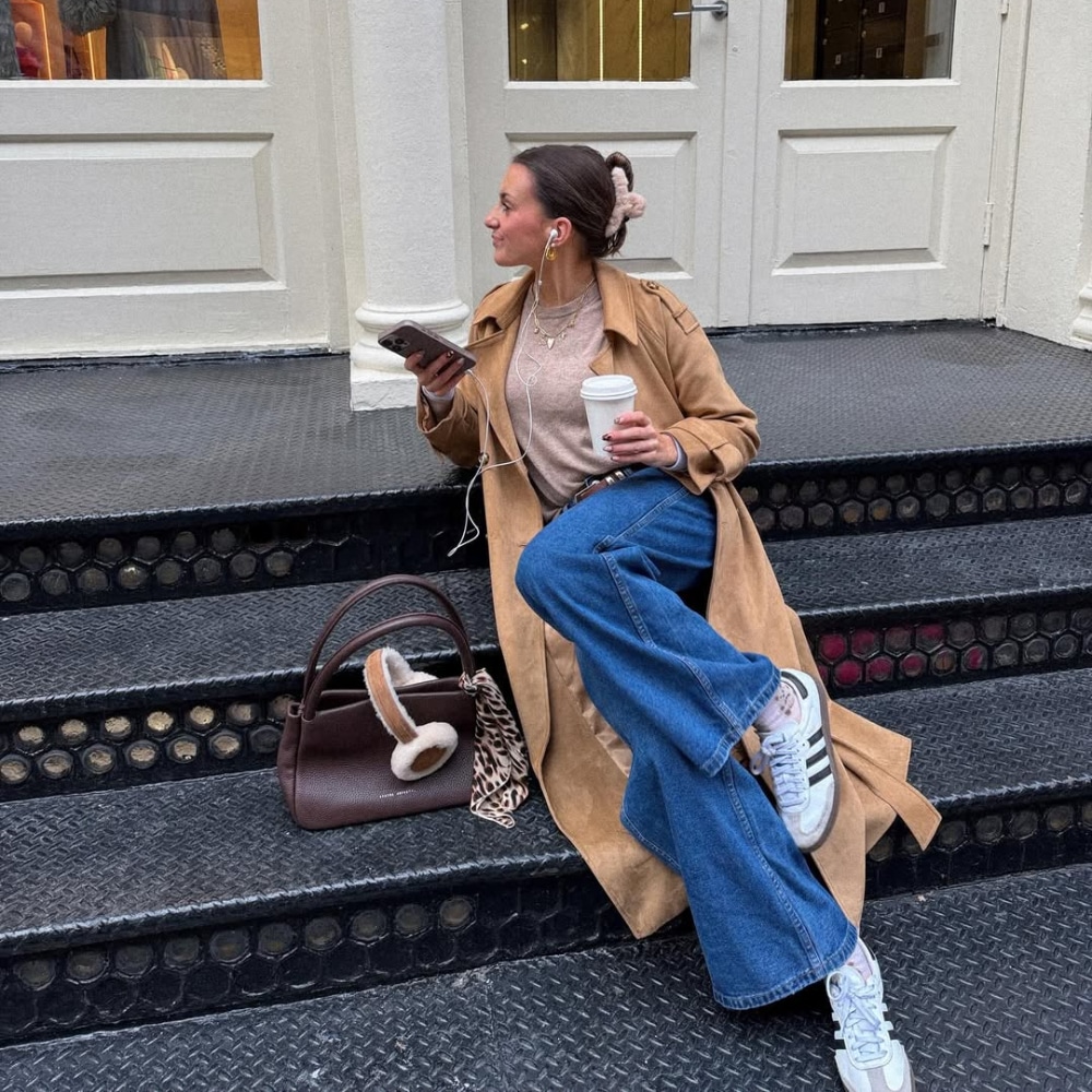 A woman in a tan trench coat and blue jeans sits on black steps, holding a coffee cup and phone, with earphones in. A brown handbag with earmuffs is beside her. She is looking to her left and smiling.