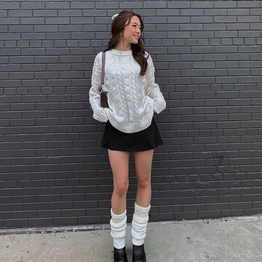A woman stands in front of a gray brick wall, wearing a white cable-knit sweater, a black skirt, black platform shoes, white leg warmers, and carrying a brown shoulder bag. She is smiling and looking to the side.
