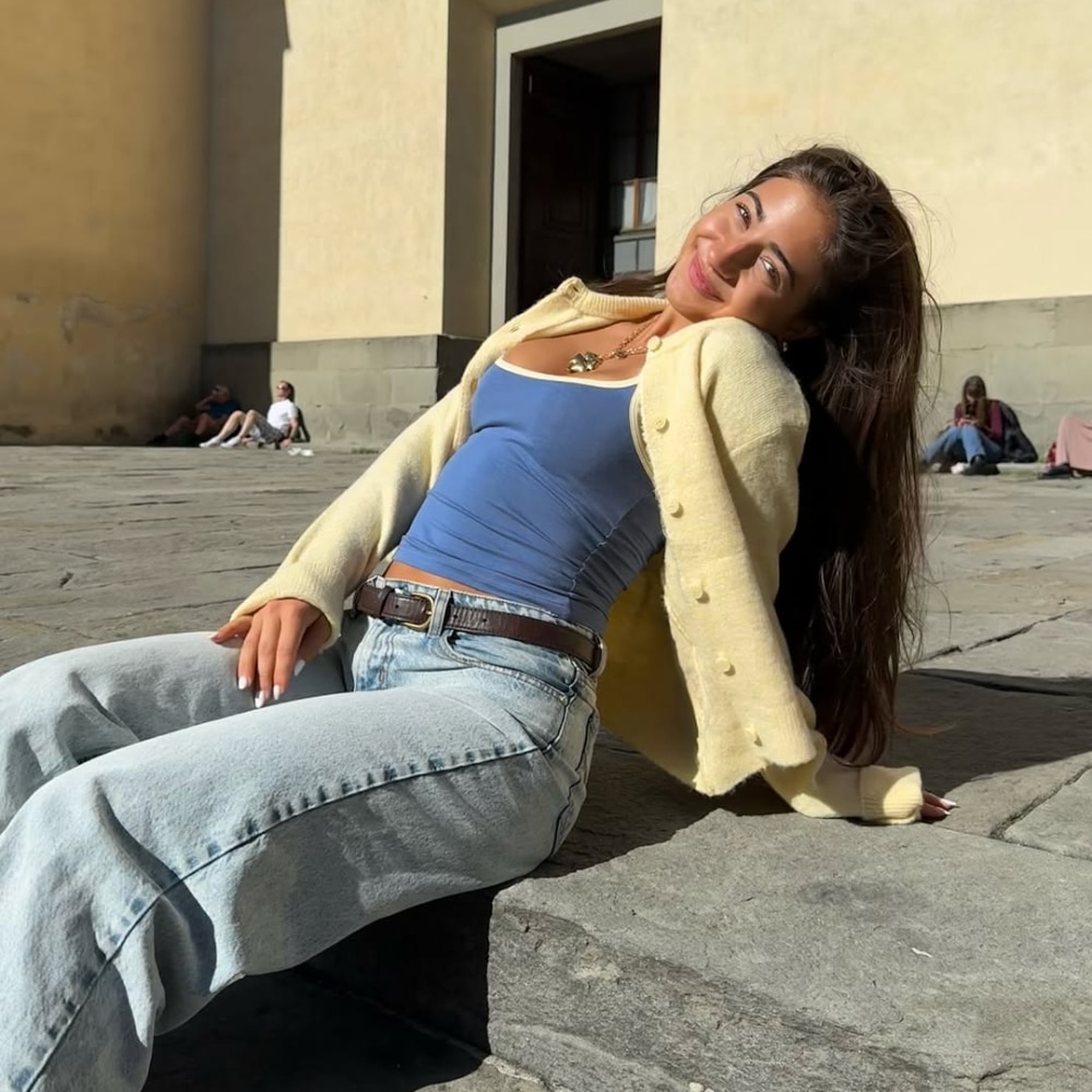 A woman with long brown hair sits on a sunlit stone pavement, leaning back on her arms and smiling. She wears a yellow cardigan, blue tank top, light blue jeans, and a brown belt. A building and people are in the background.