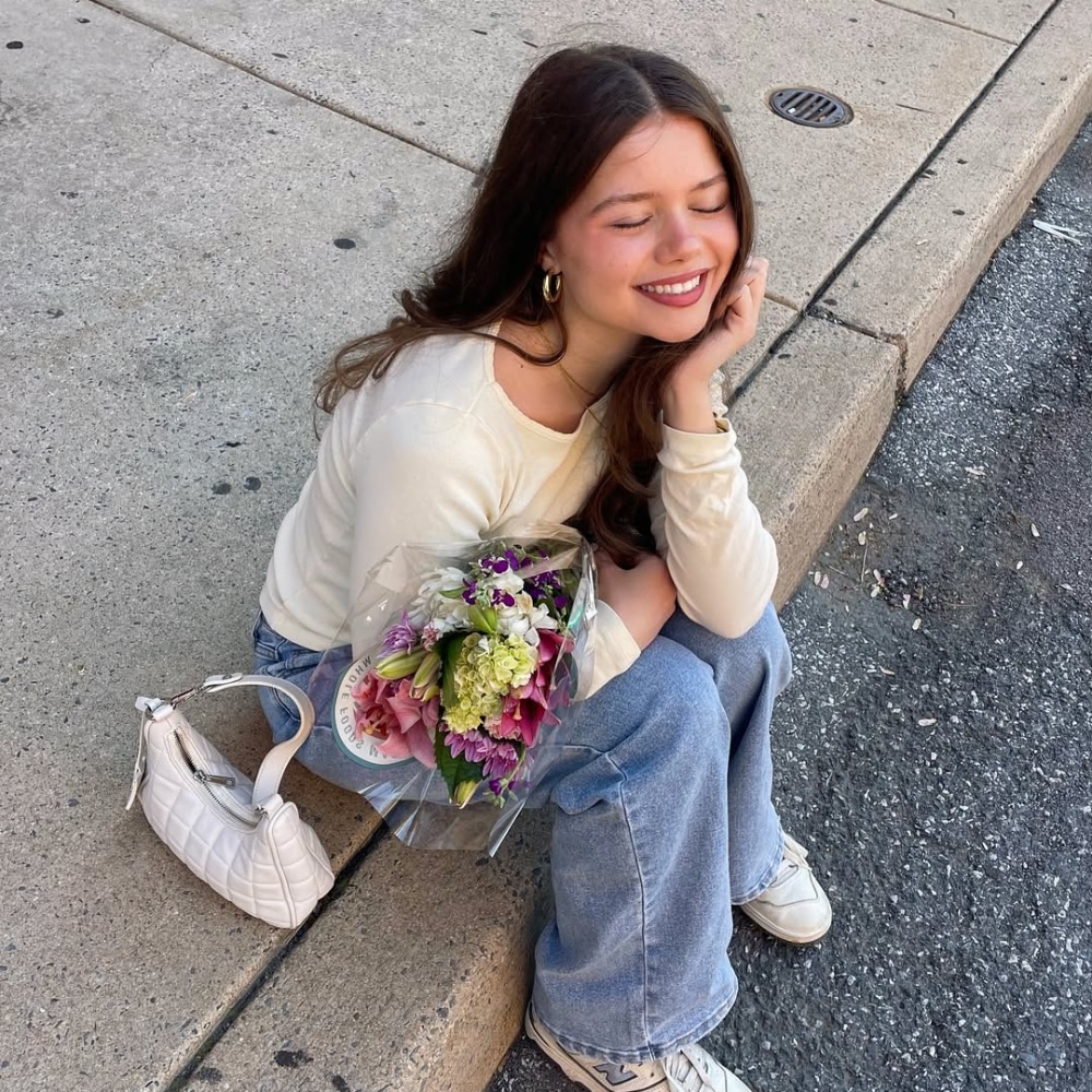 A young woman with long brown hair sits on a sidewalk curb, smiling with eyes closed. She holds a bouquet of flowers and rests her chin on her hand. She wears a cream top, blue jeans, white sneakers, and has a white purse beside her.