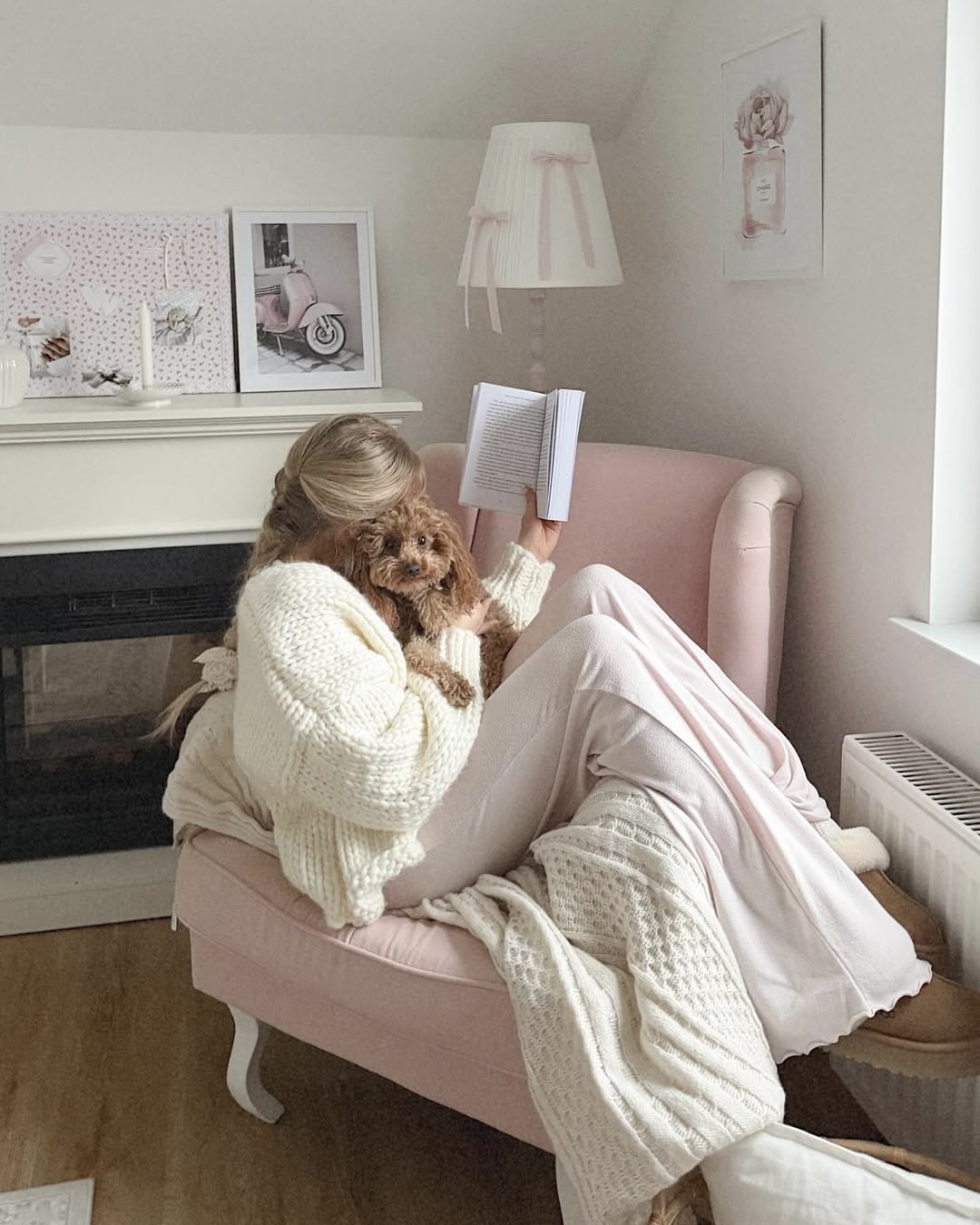 A woman in a cozy white sweater lounges on a pink chair, reading a book about coping with homesickness, with a small brown dog on her lap. The softly decorated room in white and pink offers comfort with framed art, a lamp, and a blanket.