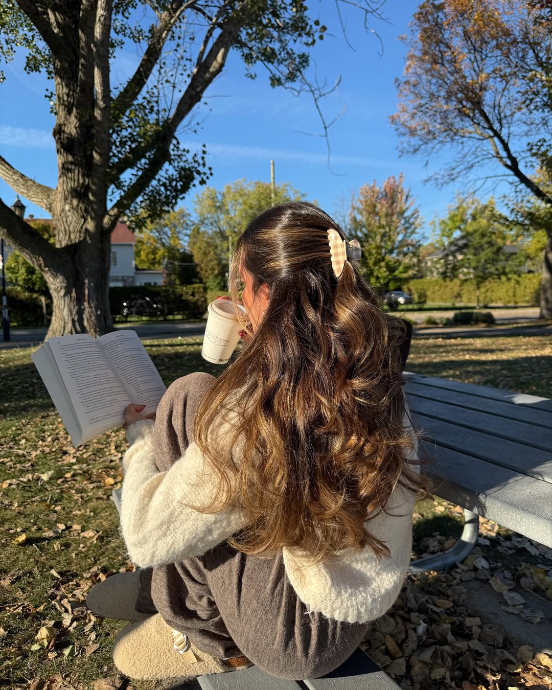 A woman with long wavy hair sits on a bench outdoors, reading a book and holding a drink. She wears cozy clothes and slippers, embracing comfort while dealing with homesickness. Autumn leaves and clear blue sky create a peaceful backdrop.
