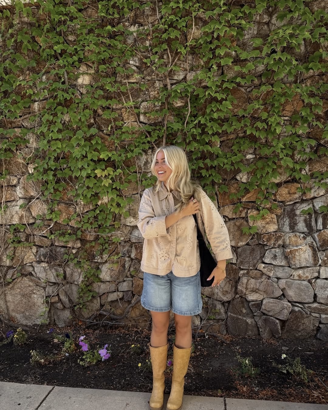 A woman with blonde hair stands smiling in front of a stone wall covered with green ivy, her relaxed pose suggesting she’s coping with homesickness as she enjoys nature. She wears a beige jacket, denim shorts, tan boots, and holds a black object in one hand. Flowers bloom below.