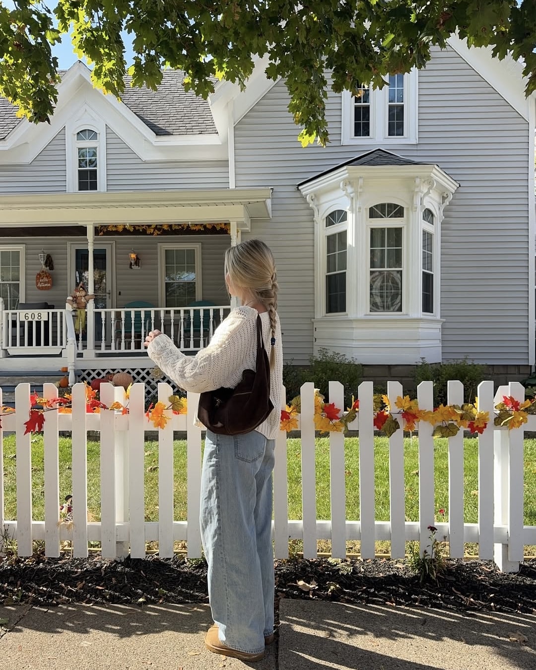 A person with braided blonde hair, wearing a cream sweater and loose jeans, stands by a white picket fence decorated with fall leaves, looking thoughtful—perhaps coping with homesickness in front of a gray house adorned for autumn.