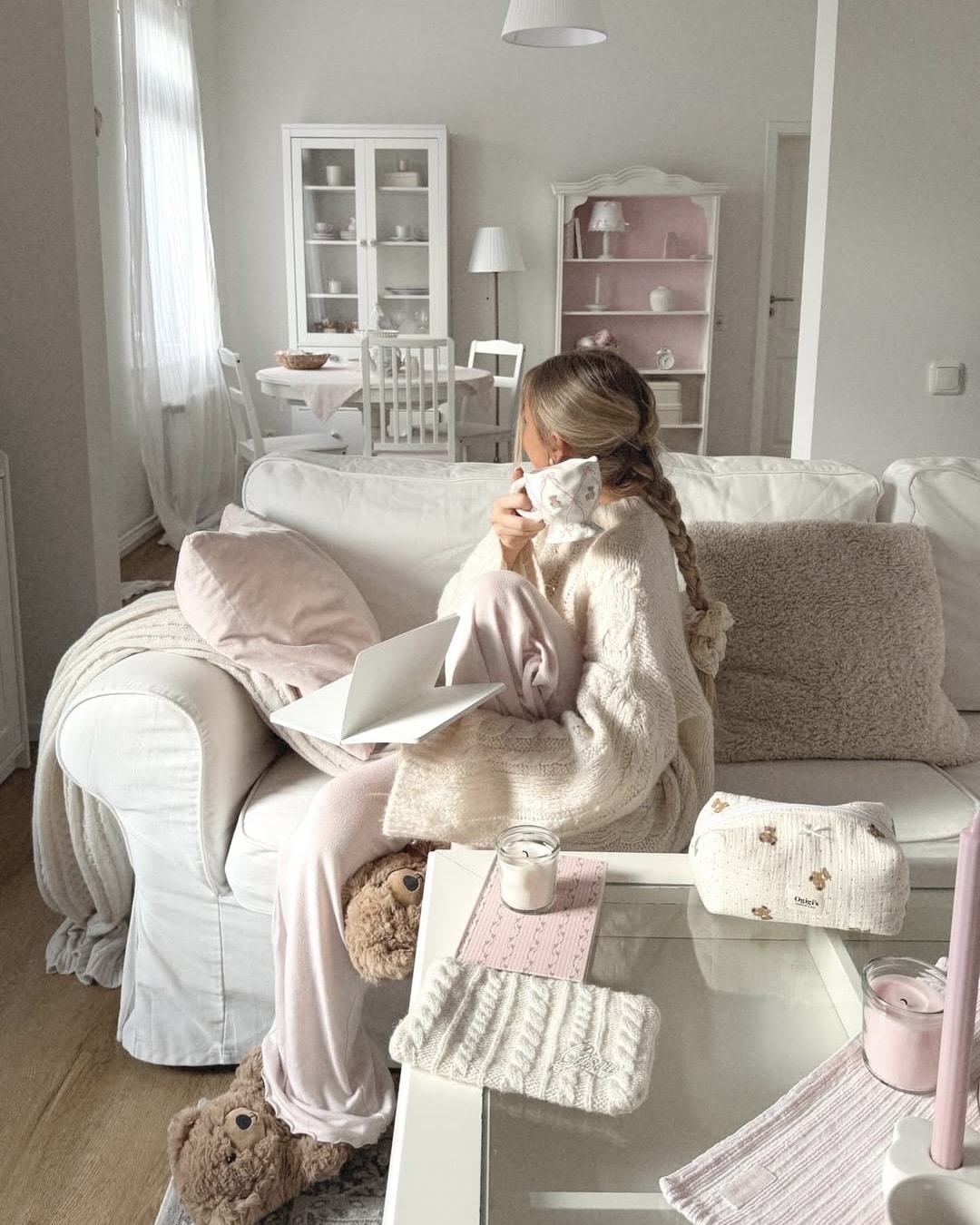 A woman coping with homesickness sits on a white sofa in a cozy, pastel-themed living room, wrapped in a blanket with a laptop on her lap. She drinks from a mug, wearing soft pajamas and bear slippers. The space is softly lit and neatly decorated.