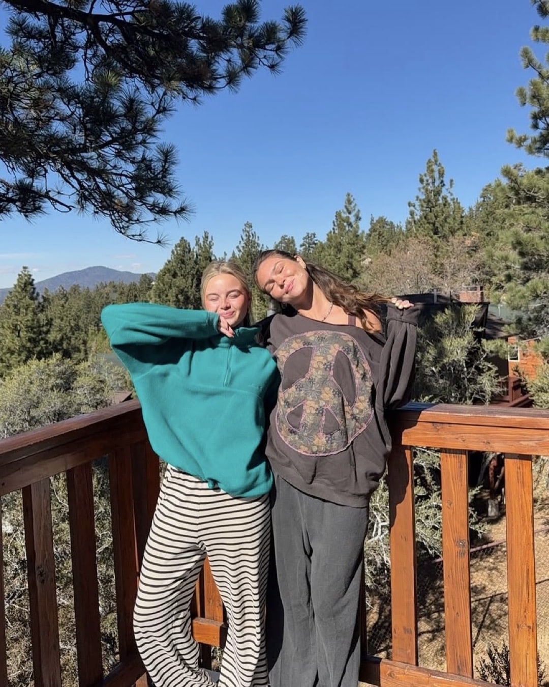 Two women, coping with homesickness, stand on a wooden deck surrounded by trees under a clear blue sky. One wears a teal sweater and striped pants; the other has a peace sign sweatshirt and gray pants. Both smile and pose casually.