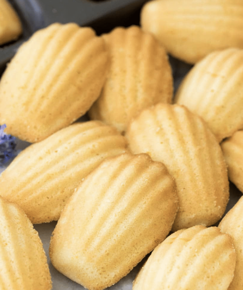 Golden madeleine cookies with a ridged shell-like pattern are closely arranged on a tray. Their lightly browned edges and soft texture are visible.
