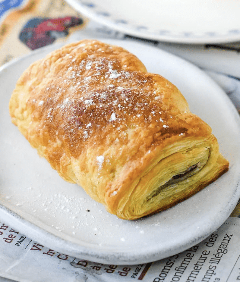 A golden, flaky pain au chocolat dusted with powdered sugar sits on a white plate, with part of a newspaper and another plate in the background.