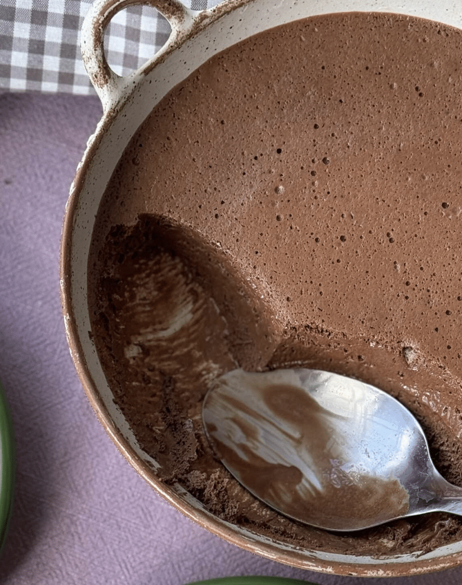 A ceramic dish of chocolate mousse with a smooth, airy texture, partially scooped out by a spoon resting inside. The background includes a purple cloth and a checked napkin.