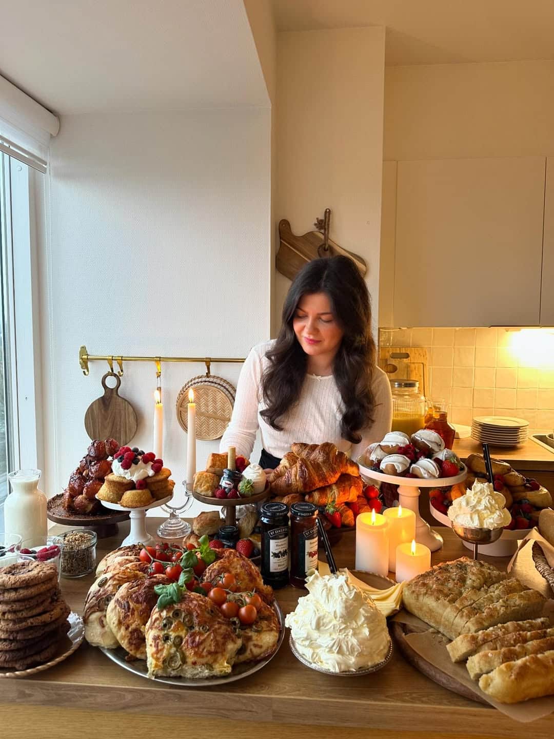 A woman with long dark hair stands behind a table filled with various pastries, breads, cakes, and desserts. The table is decorated with candles and jars of jam in a cozy, warmly lit kitchen.