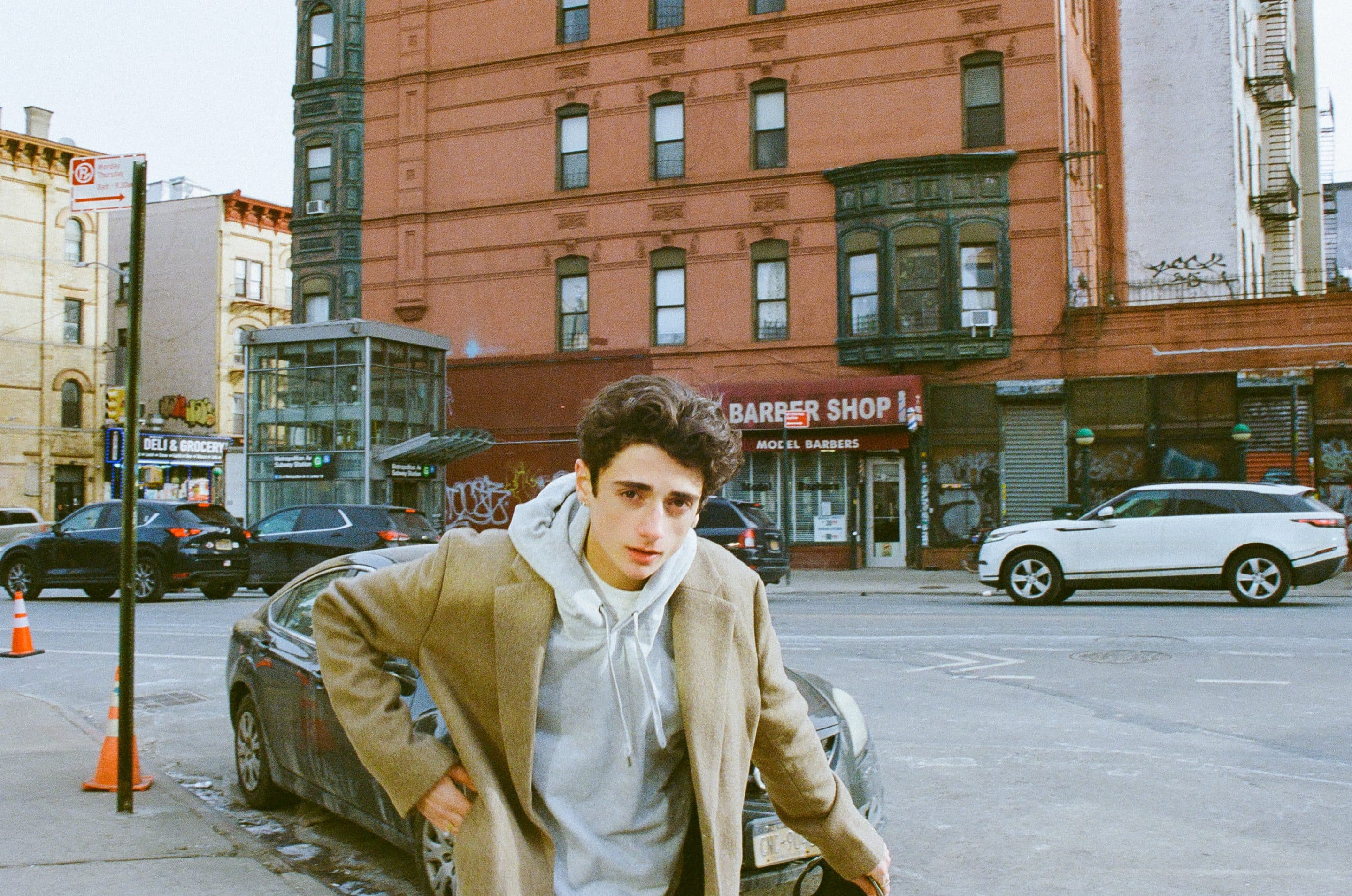 A young person in a tan coat and gray hoodie stands on a city street near parked cars and a red brick building with Dom Innarella's barbershop in the background.