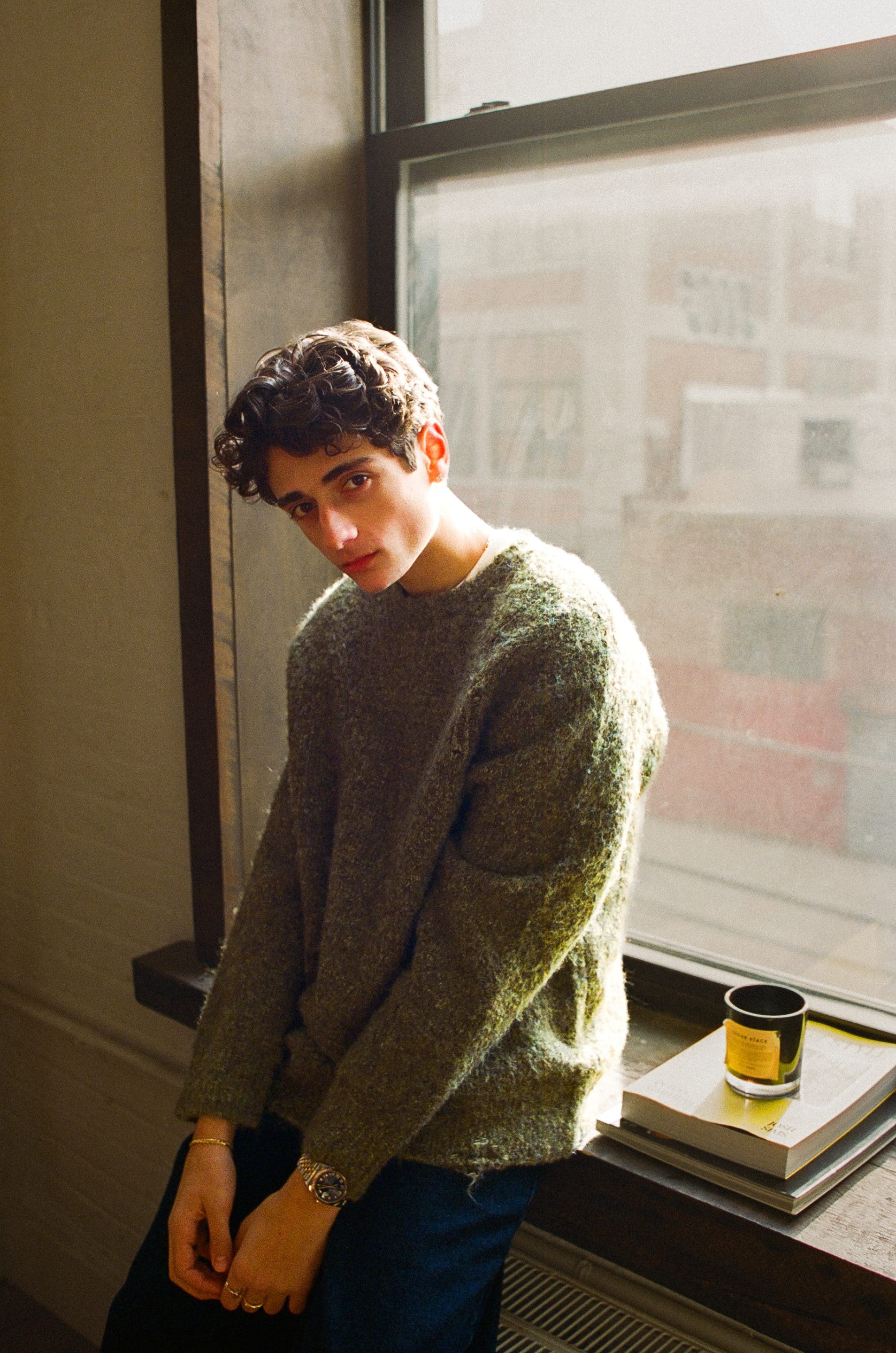 A young person with curly hair wearing a cozy green sweater sits on a windowsill, leaning forward with a relaxed expression. Sunlight streams through the window, illuminating a candle and stacked books nearby—an inviting Dom Innarella moment.