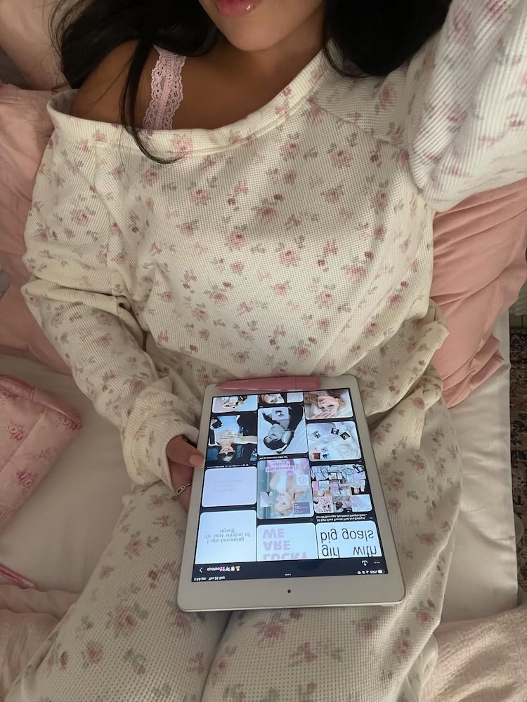 A woman in floral pajamas sits on a bed with pink pillows, holding a tablet showing a grid of images—perhaps browsing mental health tips. Only her lower face is visible, and she appears relaxed in this cozy, calming setting.
