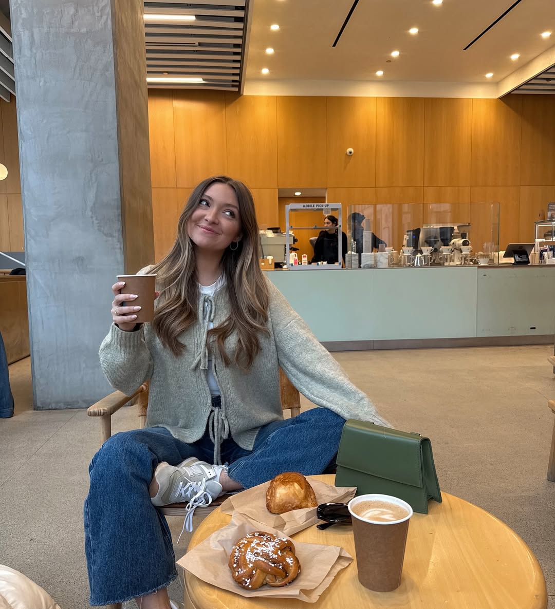 A woman with long hair sits cross-legged at a café table, smiling and holding a coffee cup. She wears a green tee, paired perfectly with her green purse, while two pastries, another coffee cup, and sunglasses sit before her. The café has wooden walls and modern decor.