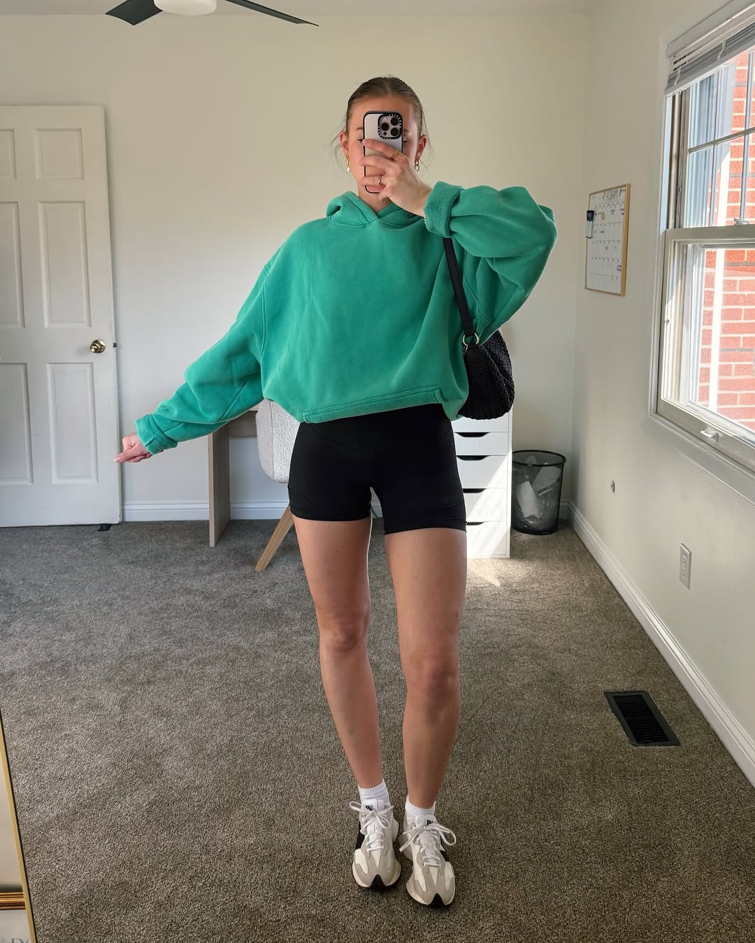 A person stands in a bright room, taking a mirror selfie in a green hoodie—perfect for St. Patrick's Day clothing—paired with black shorts, white socks, and chunky sneakers. The room has a desk, chair, window, and a whiteboard on the wall.