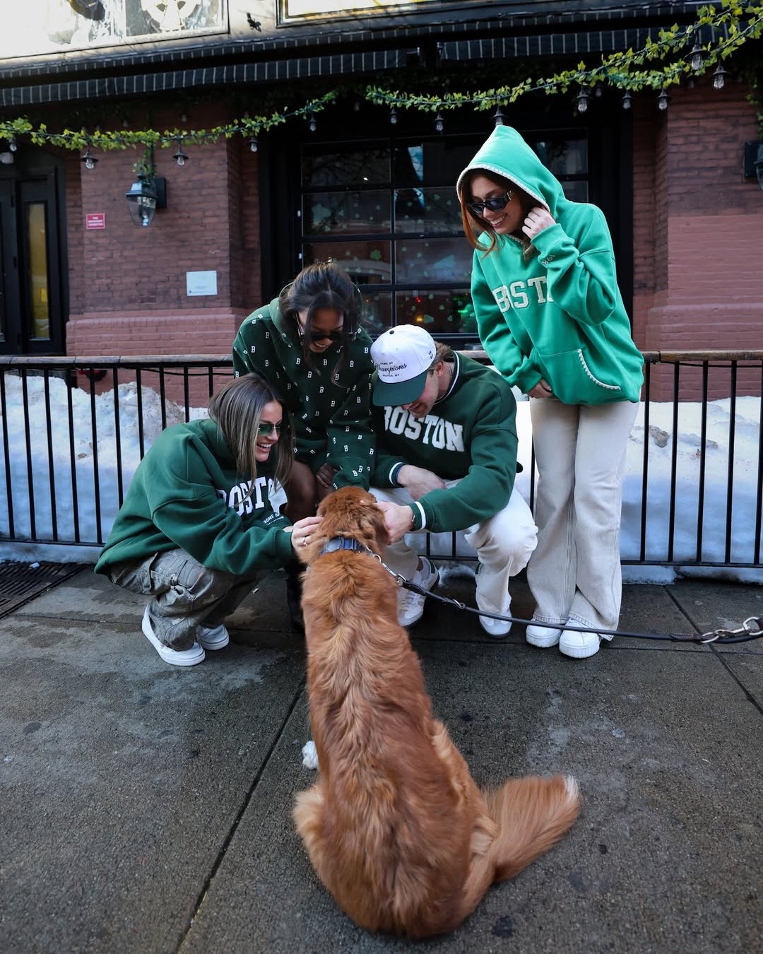 Four people in festive outfits—green Boston sweatshirts and sunglasses—are smiling and petting a golden retriever on a city sidewalk in front of a brick building with snow on the ground.