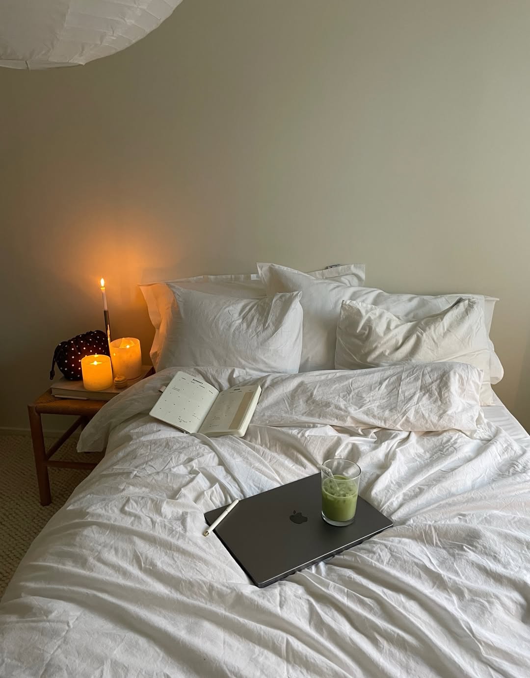 A cozy bed with white bedding sets the scene for a peaceful Sunday Reset Routine—an open notebook, closed laptop with a green drink on top, and three lit candles on a bedside table capture the calm, Spring mood in soft natural lighting.