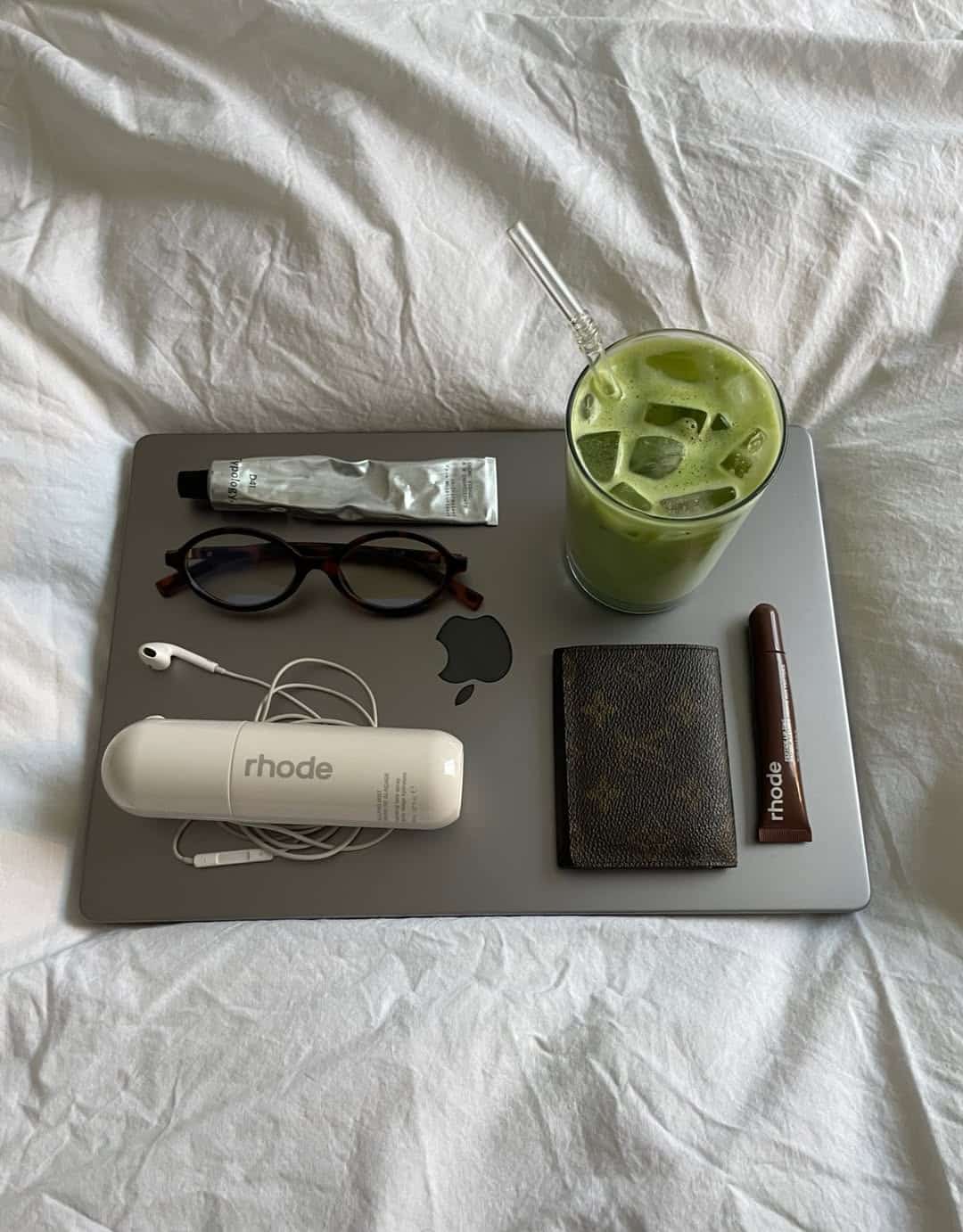 A closed MacBook on a bed holds glasses, a tube of cream, a green iced drink with a straw, a Rhode case with earbuds, a wallet, and a Rhode lip product—perfect essentials for your Spring Sunday Reset Routine.