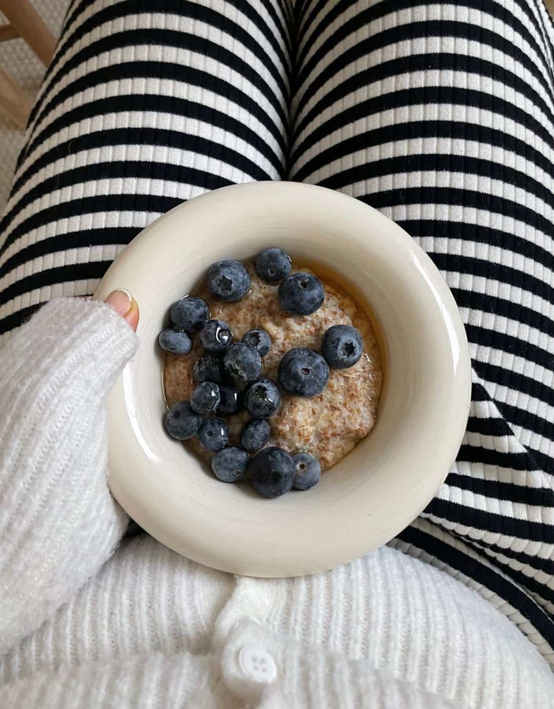 A person in striped black-and-white pants and a white sweater holds a bowl of oatmeal topped with fresh blueberries on their lap, enjoying a peaceful Sunday routine.