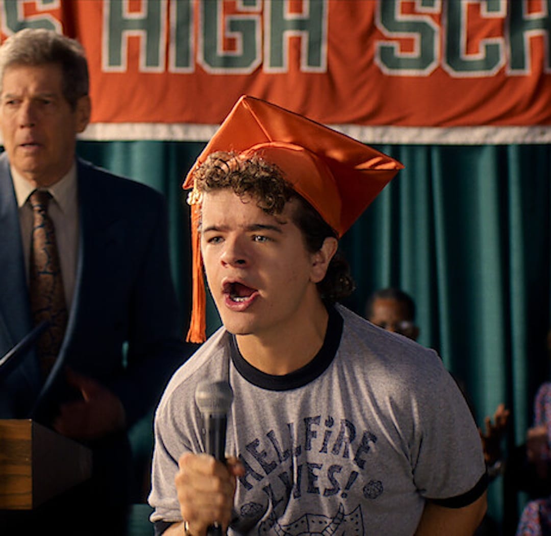 A young man with curly hair wears an orange graduation cap and holds a microphone, passionately speaking about his graduation goal. He stands in front of a banner and podium, with a man in a suit behind him.