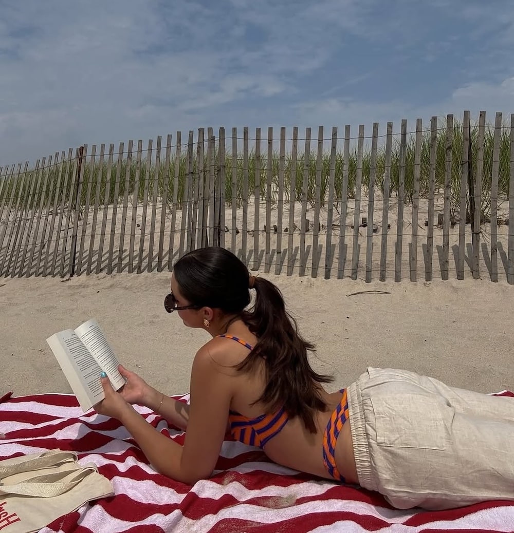 A person wearing sunglasses, a striped bikini top, and light pants lies on a red and white striped towel at the beach, reading a book from the June/July issue of Girls' Life. A wooden fence and dune grass are in the background under a partly cloudy sky.