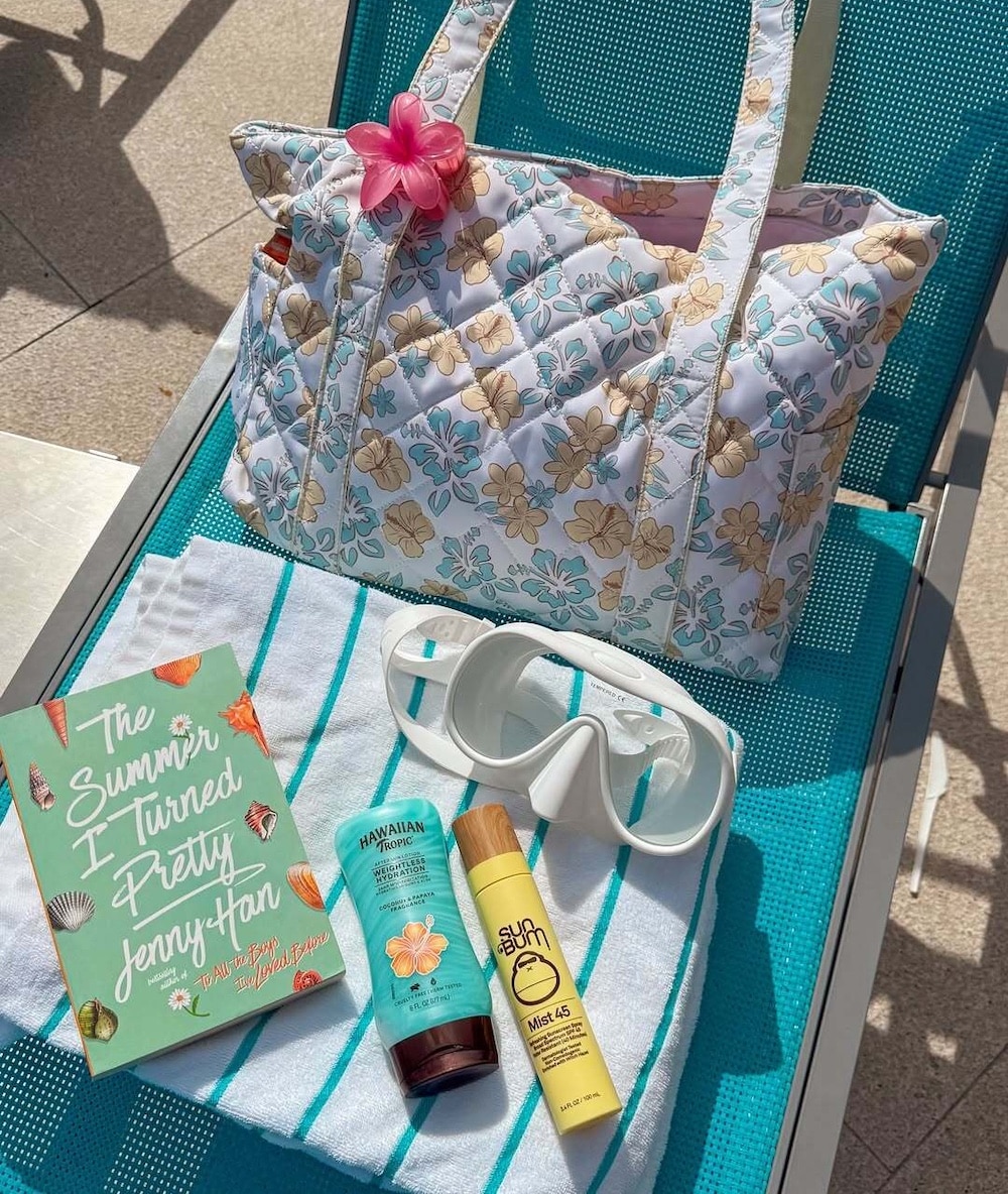 A quilted floral beach bag on a striped towel with the June/July issue of Girls' Life, The Summer I Turned Pretty, sunscreen, tanning lotion, a Sun Bum face stick, white sunglasses, and a pink flower on a turquoise lounge chair.