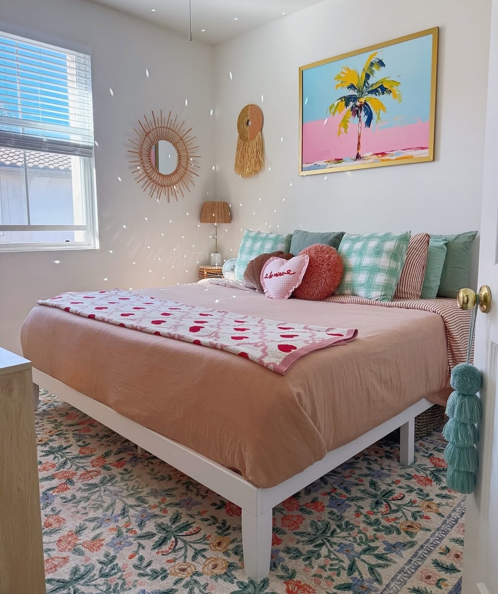 A bright bedroom with a large bed covered in pink and green bedding, a patterned rug, and colorful pillows inspired by the June/July issue of Girls' Life featuring Malia Baker. Sunlight creates sparkling reflections as decorative items hang above the bed.