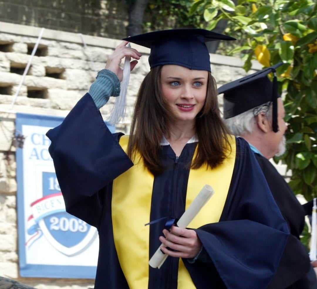 A young woman in a graduation cap and gown smiles while holding a diploma, celebrating her graduation goal. She wears a yellow stole and touches her cap, with another graduate and a school banner visible in the background.