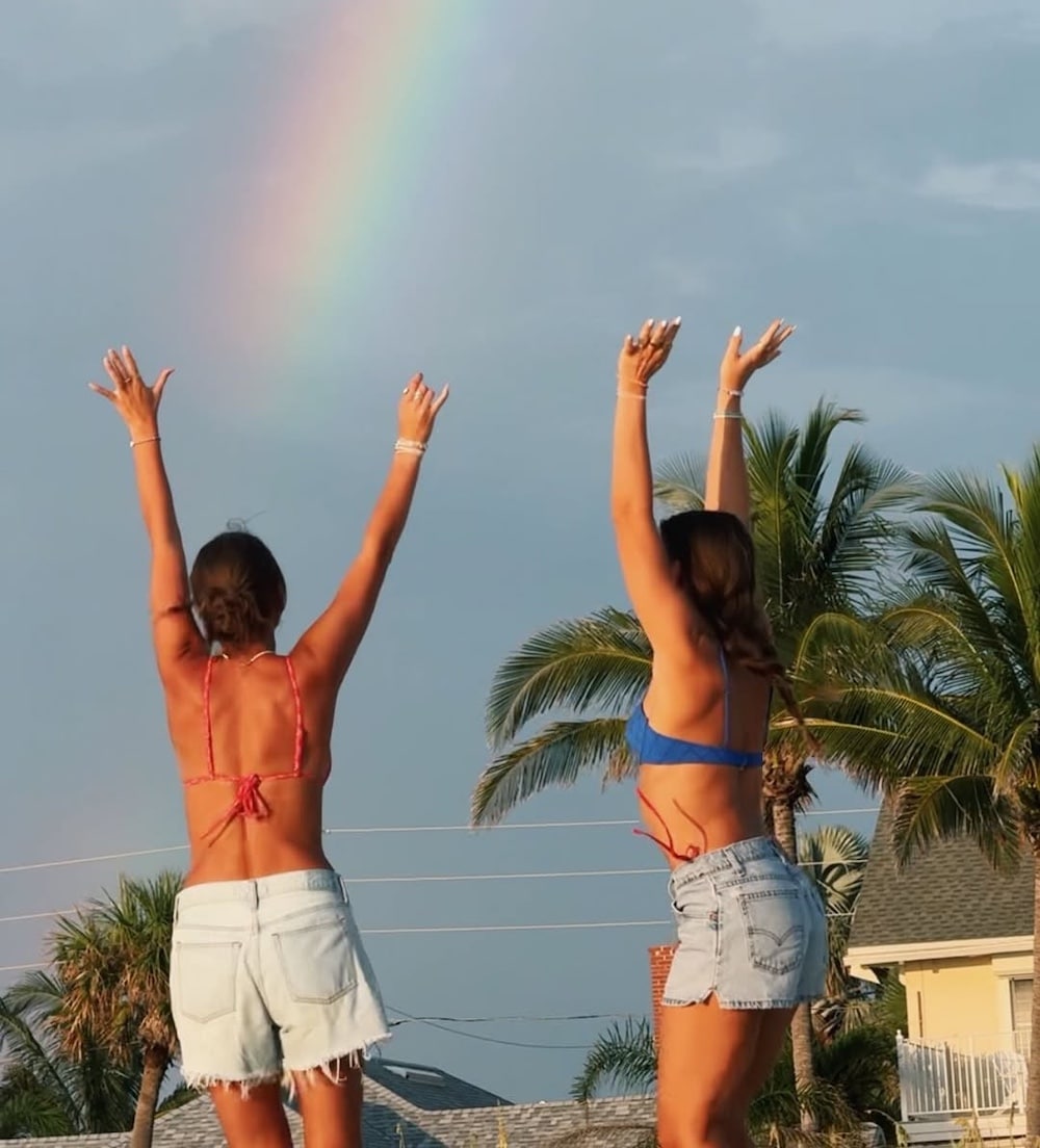 Two women in swimsuits and denim shorts stand with arms raised toward a bright rainbow, capturing the carefree vibe of a Girls' Life June/July issue cover. They're surrounded by palm trees and houses under a partly cloudy sky.
